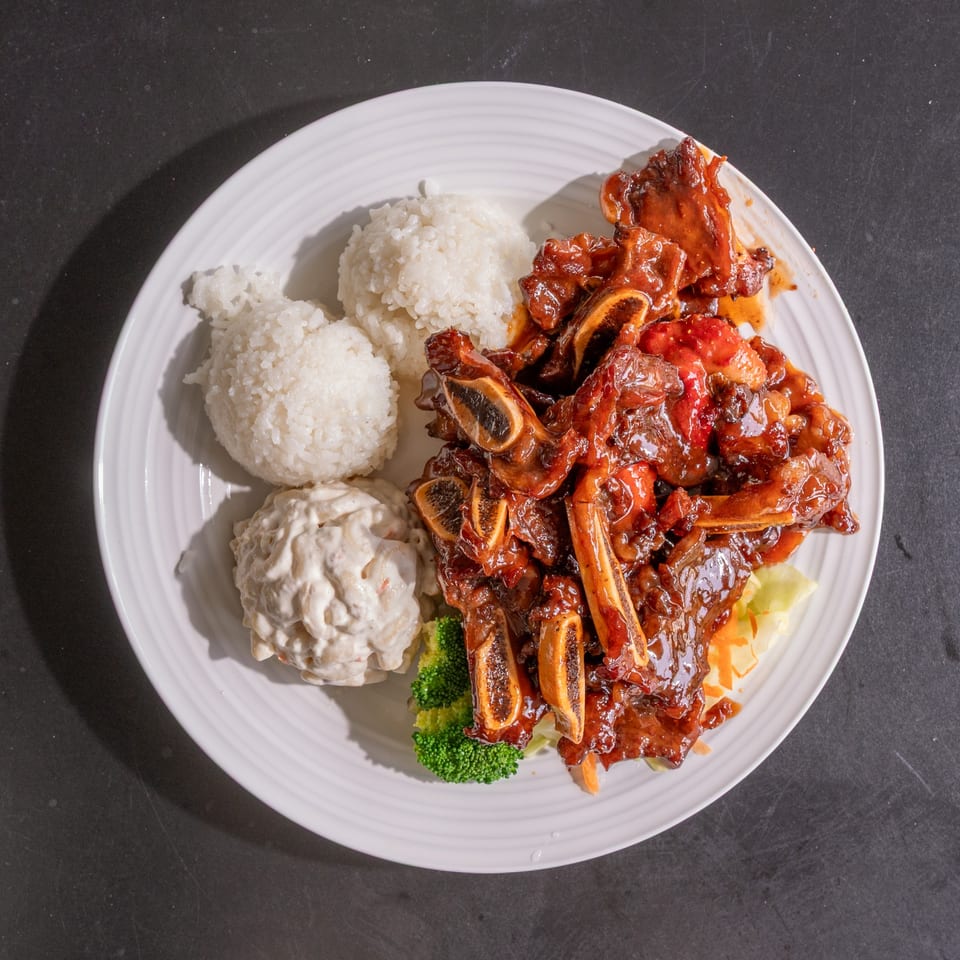A plate of BBQ short ribs with rice, macaroni salad, and broccoli on a dark background.