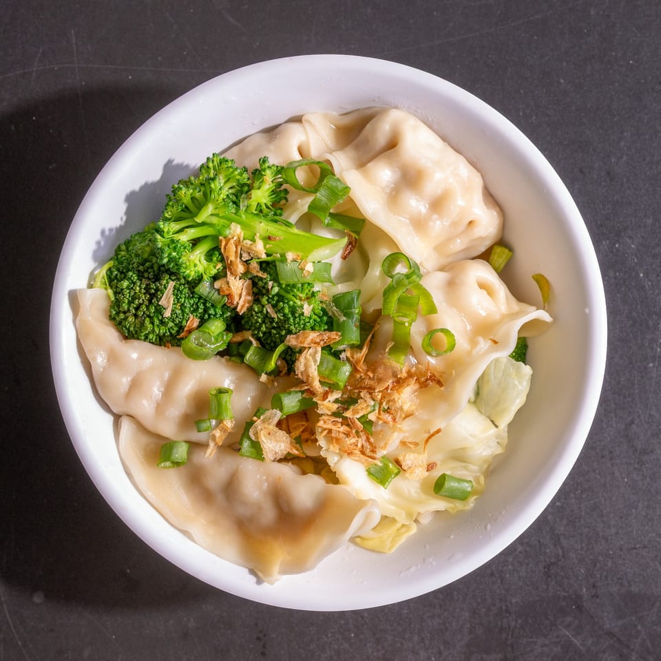 Overhead view of dumplings with broccoli, green onions, and fried shallots in a white bowl.