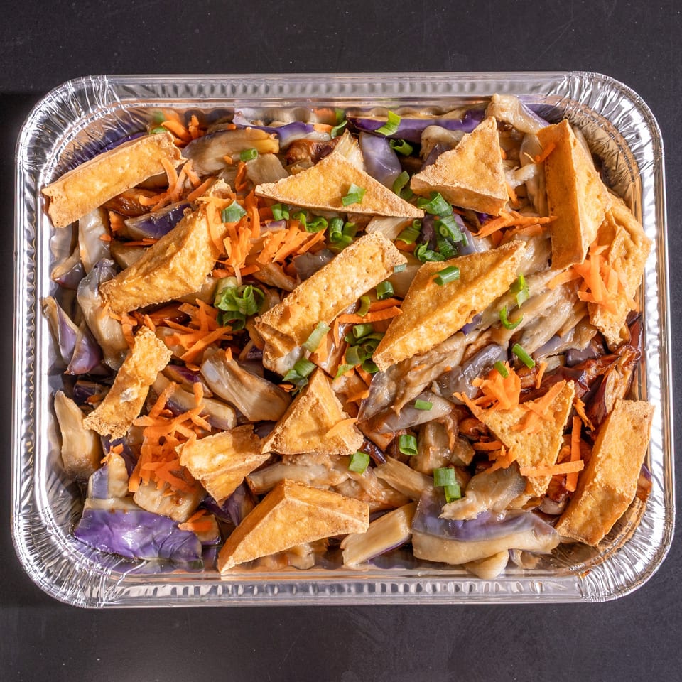 A foil tray filled with eggplant, fried tofu triangles, shredded carrots, and sliced green onions.