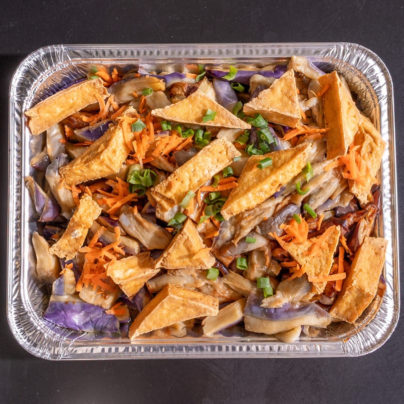 A foil tray filled with eggplant, fried tofu triangles, shredded carrots, and sliced green onions.