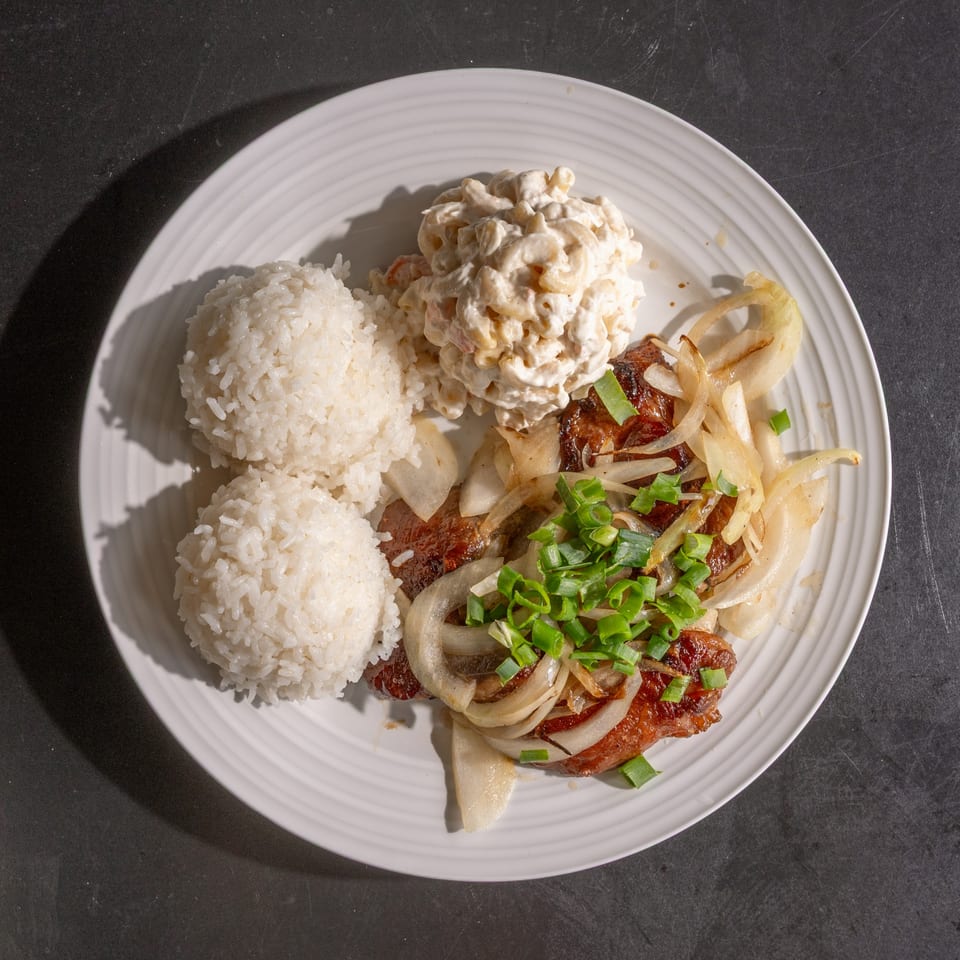 A white plate holds two scoops of rice, macaroni salad, and grilled meat with onions and scallions.